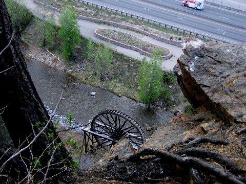 A view from above showing a riverbank with a waterwheel partially submerged in the water, surrounded by trees. In the background, a road can be seen with vehicles passing by. The scene is set in a natural landscape, with rocky terrain and greenery visible. Waterline mountain bike trail.