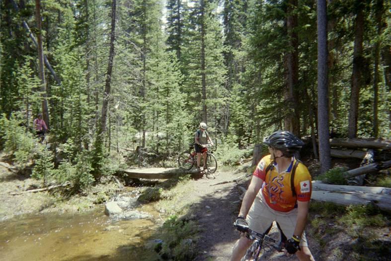 Two mountain bikers on a wooded trail near a small stream. One biker, wearing a colorful jersey, looks back while another biker in the background pauses on the path. The surrounding forest is dense with pine trees. North Lone Pine mountain bike trail.