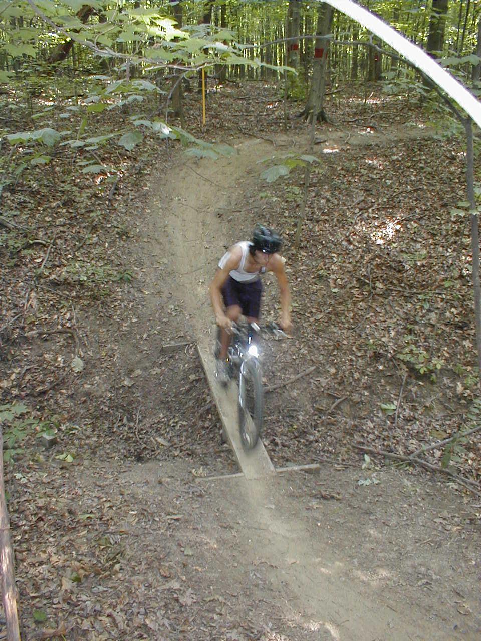 A cyclist riding a mountain bike along a narrow wooden bridge on a dirt trail in a forested area, surrounded by trees and fallen leaves. Alum Creek Phase II mountain bike trail.
