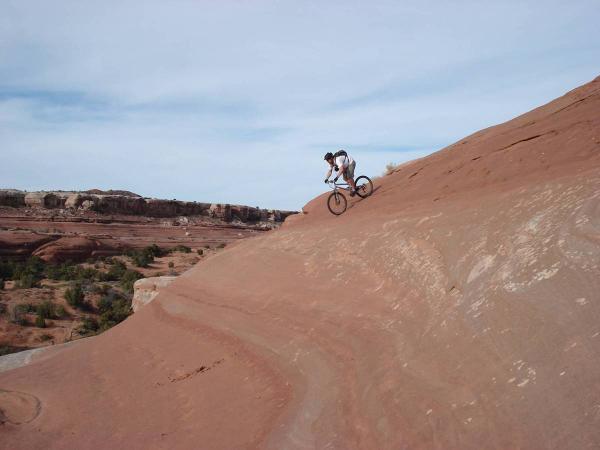 A mountain biker riding down a steep, rocky slope in a desert landscape, showcasing the rider's skill and the natural red rock formations in the background under a clear sky. Bartlett Wash mountain bike trail.