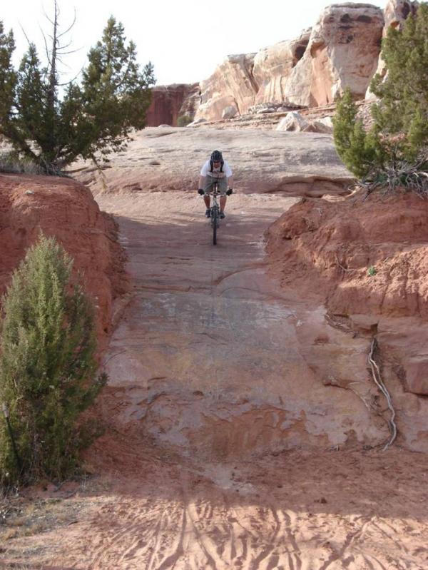 A mountain biker rides down a rocky trail, surrounded by reddish terrain and sparse greenery. The path is steep and uneven, leading towards a rugged landscape with cliffs in the background. Bartlett Wash mountain bike trail.
