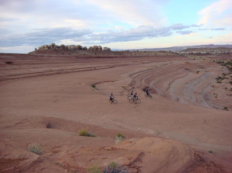 Three mountain bikers ride along the textured red rock terrain under a partly cloudy sky. In the background, a rock formation rises above the landscape, while the foreground features layered rock patterns and sparse vegetation. Bartlett Wash mountain bike trail.