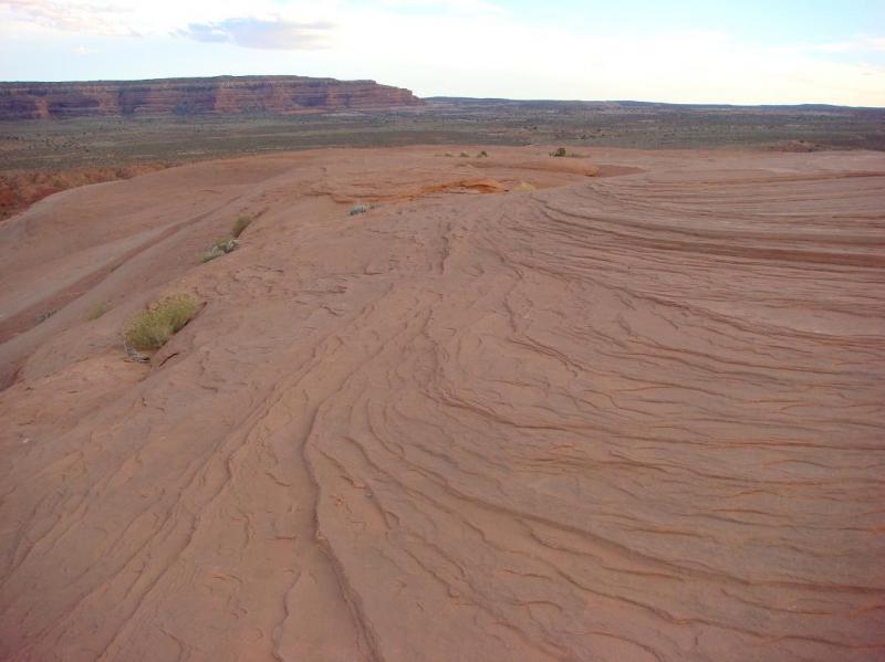 Desert landscape featuring textured reddish-brown rock formations and gentle slopes, with distant mesas and a blue sky in the background. Sparse vegetation is visible among the rock layers. Bartlett Wash mountain bike trail.