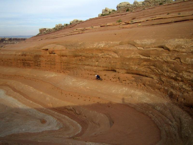 A person riding a mountain bike along a layered sandstone landscape, showcasing various shades of red and brown rock formations under a partly cloudy sky. Bartlett Wash mountain bike trail.