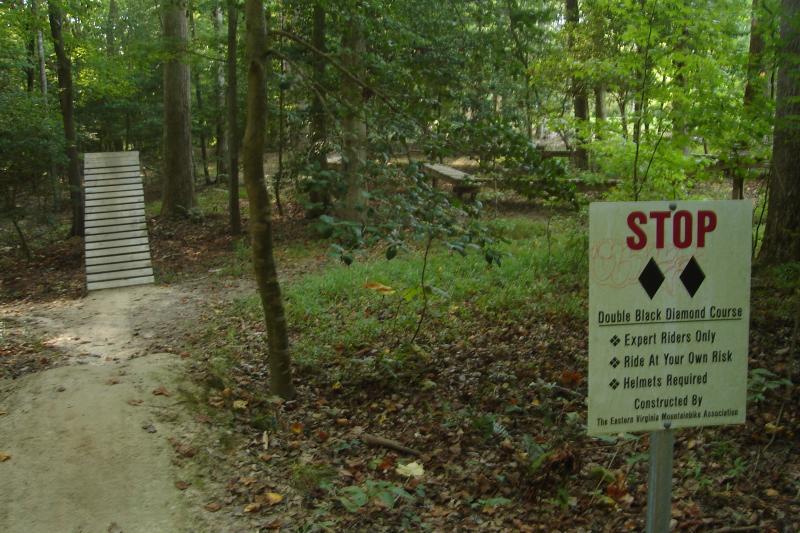 A wooded area featuring a dirt path leading to a wooden ramp. A sign labeled "STOP" indicates the start of a double black diamond mountain biking course, warning that it is for expert riders only, and helmets are required. The surroundings are lush with greenery and foliage. New Quarter Park mountain bike trail.