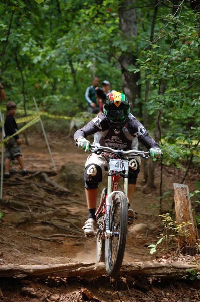 A mountain biker navigating a wooded trail, jumping over a log while wearing protective gear, including a helmet and knee pads. In the background, a few spectators watch the action. The scene captures the excitement of downhill biking in a natural outdoor setting. Paris Mountain State Park mountain bike trail.
