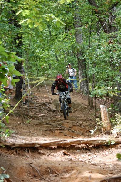 A mountain biker navigates a rugged trail surrounded by trees, wearing a helmet and protective gear. In the background, a spectator watches, and yellow tape marks the course boundaries. The uneven terrain features exposed roots and dirt, indicative of a challenging biking path. Paris Mountain State Park mountain bike trail.