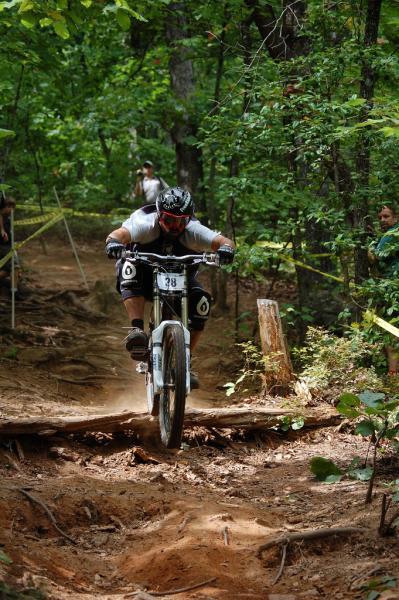 A mountain biker in a black helmet and protective gear navigates a dirt trail in a wooded area, leaping over a log while kicking up dust. Spectators can be seen along the trail, surrounded by lush green trees. Paris Mountain State Park mountain bike trail.