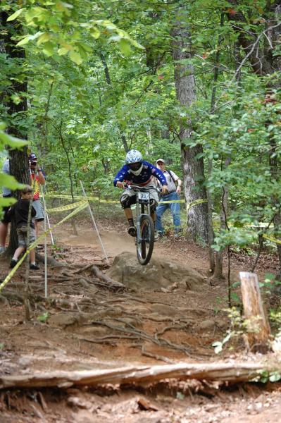 A mountain biker in protective gear is captured mid-air as they jump over a large rock on a forested trail. Surrounding trees provide a vibrant green backdrop, while spectators can be seen in the distance observing the ride. Yellow caution tape lines the track, indicating the race area. Paris Mountain State Park mountain bike trail.