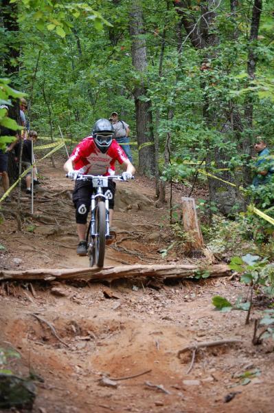 A mountain biker in a red jersey and black shorts navigates a dirt trail, skillfully maneuvering over a wooden log obstacle. Surrounded by lush green trees, onlookers can be seen in the background, observing the race. The scene captures the excitement of downhill biking in a natural setting. Paris Mountain State Park mountain bike trail.
