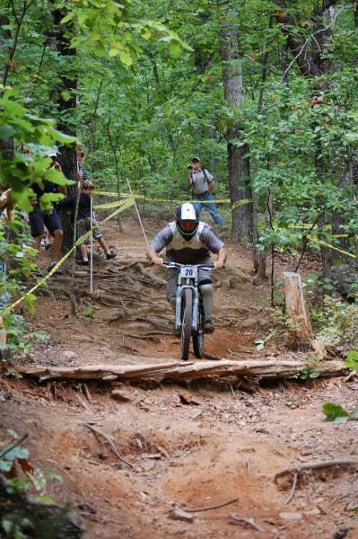 A mountain biker navigating a rugged trail, focused on the path ahead as they approach a wooden bridge. Surrounded by lush greenery and trees, a few spectators can be seen in the background observing the ride. Paris Mountain State Park mountain bike trail.