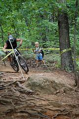 A mountain biker navigating a challenging trail surrounded by trees, while a child watches nearby. The scene includes visible roots and a large rock on the path, along with caution tape marking the area. Paris Mountain State Park mountain bike trail.