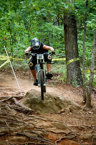 A mountain biker in protective gear is airborne as they jump off a rock on a forested trail, surrounded by trees and yellow caution tape. The rider is wearing a black helmet, jersey, and knee pads, with the number 34 displayed on their shorts. Paris Mountain State Park mountain bike trail.