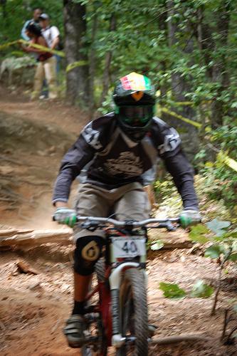 Action shot of a mountain biker tackling a dirt trail in a forested area. The rider, wearing a helmet and protective gear, is navigating over a log while surrounded by trees. In the background, a second person can be seen watching the ride. The scene captures the excitement and intensity of downhill biking. Paris Mountain State Park mountain bike trail.