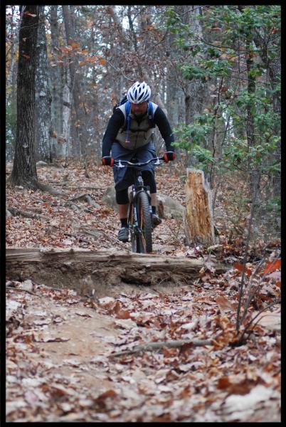 A mountain biker navigating a rugged trail, wearing a helmet and riding gear, with autumn leaves scattered on the ground and trees in the background. Paris Mountain State Park mountain bike trail.