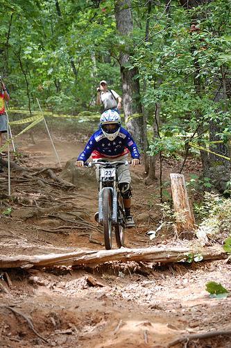 A mountain biker wearing a helmet and protective gear rides down a rugged trail surrounded by trees. The path is dusty and includes visible roots and obstacles. In the background, a person is observing the rider. The biker's jersey features a patriotic color scheme, and they are navigating the challenging terrain during a cycling event. Paris Mountain State Park mountain bike trail.