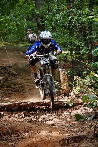 A cyclist in protective gear and a helmet is mid-jump while riding a mountain bike on a dirt trail in a wooded area. The rider wears a blue jersey with white stars and displays a race number. In the background, spectators watch the event amidst the greenery. Paris Mountain State Park mountain bike trail.