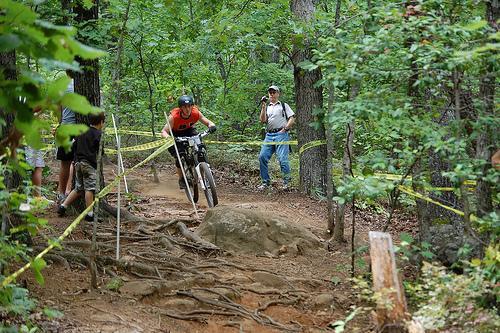 A mountain biker in an orange shirt navigates a dirt trail surrounded by trees, while two spectators observe the ride from the side. The path features exposed roots and a large rock, with yellow tape marking the course. Paris Mountain State Park mountain bike trail.