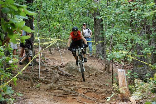 A mountain biker in a red shirt navigates a rocky trail surrounded by trees, while spectators watch from the side. Yellow caution tape lines the course, indicating the boundaries of the biking path. Paris Mountain State Park mountain bike trail.