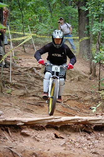A mountain biker in protective gear rides down a rocky trail in a wooded area, navigating a log on the ground. The cyclist is wearing a helmet and jersey, and has a race number 13 displayed on their bike. In the background, a spectator watches from a distance. Yellow caution tape marks the course. Paris Mountain State Park mountain bike trail.