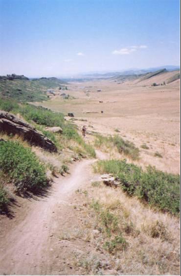 A sunlit trail winding through a grassy landscape, with rolling hills in the background and a clear blue sky. A distant figure walks along the path, surrounded by shrubs and rocky formations. Blue Sky mountain bike trail.