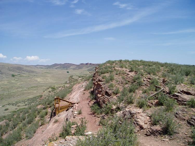 A rocky landscape with a winding dirt path leading to a viewpoint. The foreground features a rocky outcrop covered in green shrubs, while a wooden structure is positioned near the edge. In the background, rolling hills and a clear blue sky with wispy clouds are visible, creating a serene natural scene. Blue Sky mountain bike trail.