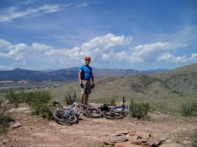 A mountain biker standing on rocky terrain, wearing a blue jersey, shorts, and a helmet, with two mountain bikes nearby. The backdrop features rolling hills and a blue sky with fluffy clouds. Blue Sky mountain bike trail.