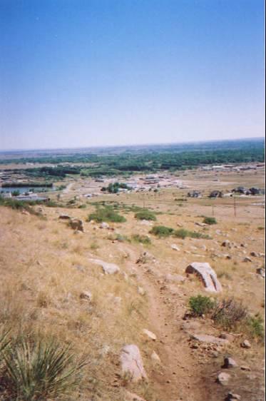 A scenic landscape viewed from a hill, showing a dirt path leading downwards. The foreground features dry grass and large rocks, while the background reveals a wide valley with a small town, fields, and trees under a clear blue sky. Foothills Trail mountain bike trail.