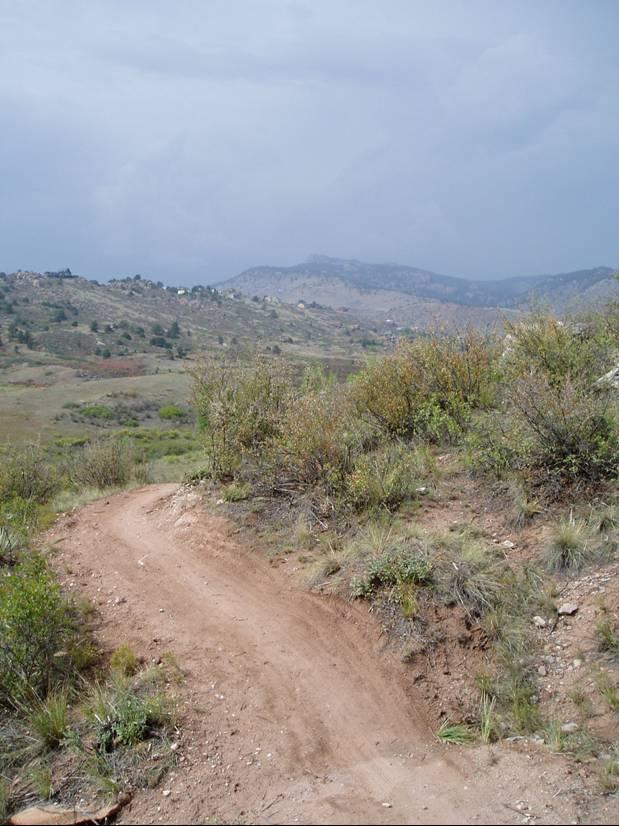 A dirt biking trail winding through a hilly landscape, surrounded by patches of green vegetation and small shrubs. The sky is overcast with gray clouds, indicating a possibility of rain. In the distance, rolling hills and a few houses can be seen against the backdrop of the mountains. Blue Sky mountain bike trail.