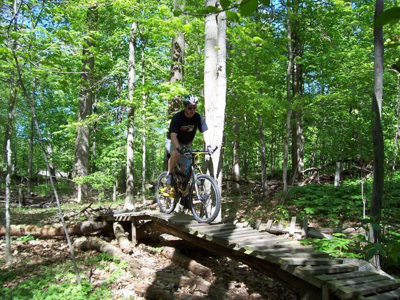 A mountain biker navigating a wooden bridge in a lush green forest, surrounded by tall trees and vibrant foliage. Sunlight filters through the leaves, creating a bright and inviting outdoor scene. Burchfield mountain bike trail.