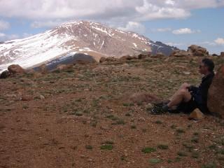 A hiker sitting on a rock in a mountainous landscape, with a snow-capped peak in the background and a blue sky dotted with clouds. The terrain is rocky and covered with sparse vegetation. Mount Baldy mountain bike trail.