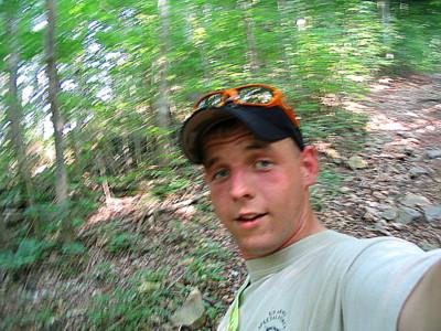 A young man taking a selfie while hiking on a rocky trail surrounded by lush green trees. The image appears slightly blurred, suggesting movement. He is wearing a cap and sunglasses. Knights Path mountain bike trail.