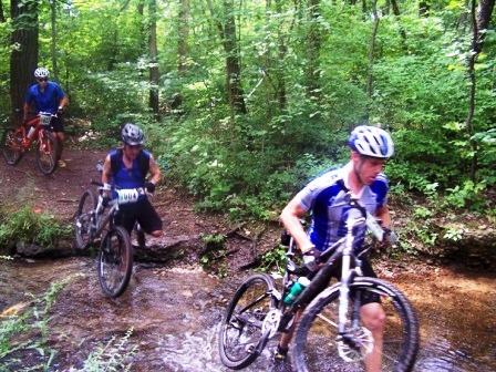 A group of mountain bikers navigating a stream in a forested area. Two riders are walking their bikes through the shallow water, while another cyclist is in the background on the trail, surrounded by lush greenery and trees. France Park mountain bike trail.