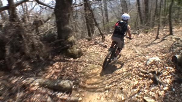A person riding a mountain bike on a dirt trail through a wooded area, surrounded by trees and scattered leaves. The rider is wearing a helmet and a backpack, navigating a path with rocks and natural terrain. Black Mountain mountain bike trail.
