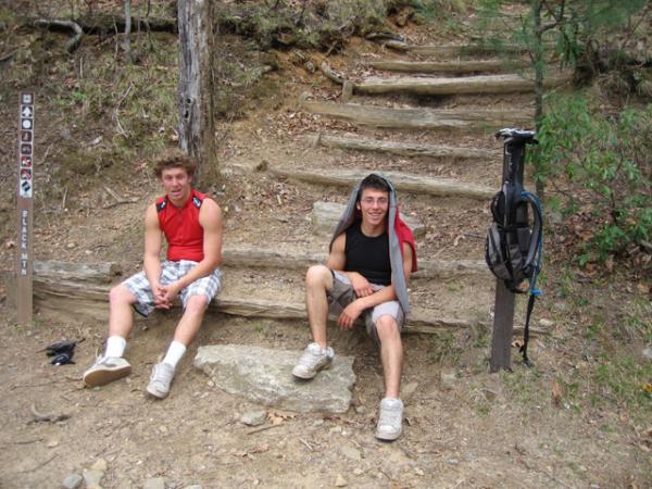 Two young men sitting on steps at a hiking trail. One is wearing a red sleeveless shirt and plaid shorts, while the other is in a black tank top with a gray hoodie draped over his head. Nearby, there is a mountain bike and trail signage in the background, surrounded by trees and earthy terrain. Black Mountain mountain bike trail.
