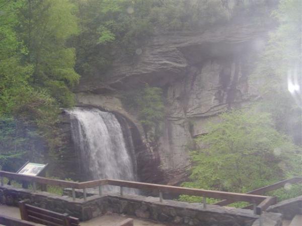 A waterfall cascading over a rocky cliff, surrounded by lush green trees. In the foreground, there is a wooden railing and a stone path, along with a signboard indicating information about the site. The scene conveys a serene natural setting. Black Mountain mountain bike trail.