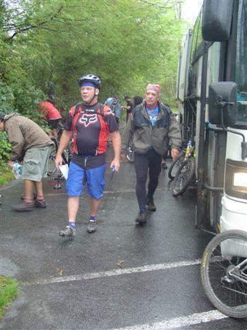 A group of mountain bikers disembarks from a bus on a rainy day, with some riders preparing their bikes while others walk along a wet road surrounded by greenery. Black Mountain mountain bike trail.