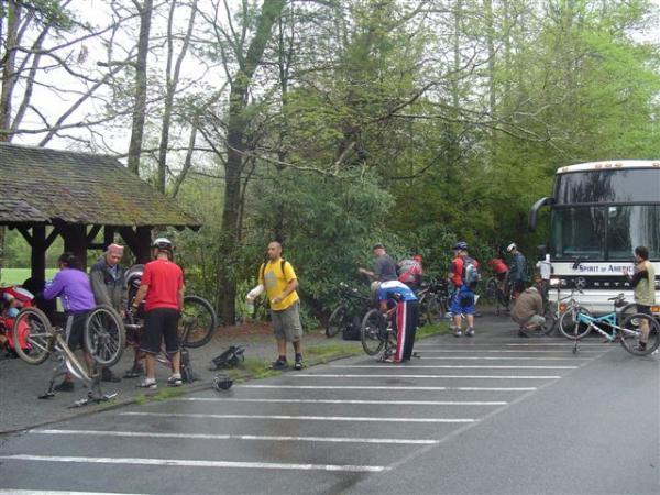 A group of cyclists gathered at a parking area near a wooded area. Some individuals are repairing bicycles, while others are adjusting gear or chatting. In the background, a bus labeled "Spirit of America" is parked next to a line of bicycles. The scene is set in a lush, green environment, indicating a cool, possibly rainy day. Black Mountain mountain bike trail.