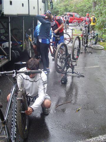 A group of cyclists working on their mountain bikes in a damp outdoor setting, with one cyclist kneeling next to a bike, adjusting the wheel, while others are seen fixing their bikes nearby. Several bicycles and vehicles are visible in the background. Black Mountain mountain bike trail.