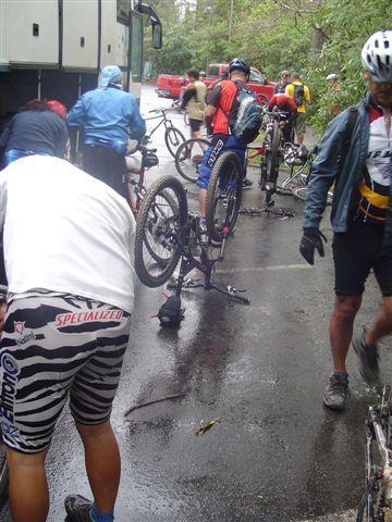 A group of cyclists is gathered on a rainy road, with several bicycles positioned upside down for repairs. Some cyclists are dressed in colorful cycling gear, and a bus can be seen in the background. The ground is wet from the rain, and the scene conveys an atmosphere of camaraderie and teamwork during a biking event. Black Mountain mountain bike trail.