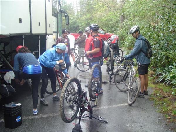 A group of mountain bikers gathered near a tour bus, preparing their bikes for a ride. Some individuals are working on bike repairs, while others are adjusting their gear. It’s a cloudy day, and the surroundings are lush with greenery, indicating a natural outdoor setting. Black Mountain mountain bike trail.