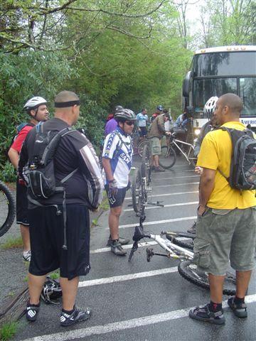 A group of mountain bikers gathered near a bus on a rainy day. Some cyclists are inspecting a bike that is on its side, while others are standing around, wearing helmets and cycling gear. Lush greenery lines the background, indicating a natural, outdoor setting. Black Mountain mountain bike trail.