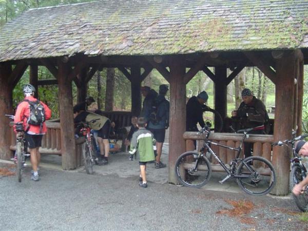 A group of people, including a child, gathered under a rustic wooden pavilion surrounded by trees. Some individuals are sitting on benches, while others are standing near their bicycles. The scene suggests a break during a biking trip, with a casually relaxed atmosphere. The weather appears overcast, possibly indicating light rain. Black Mountain mountain bike trail.