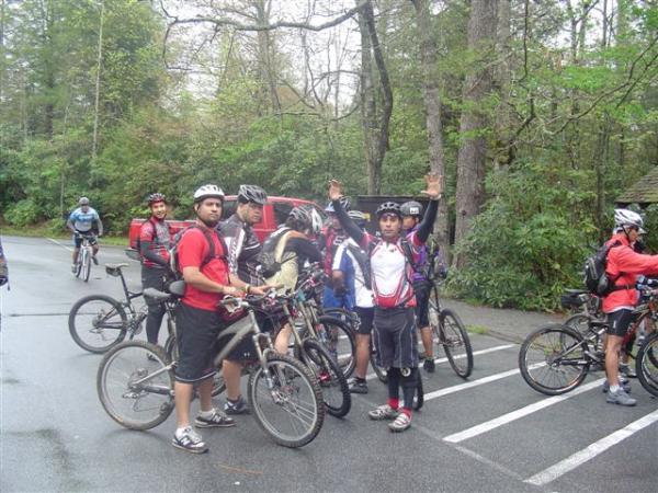 A group of mountain bikers gathered in a forested area, some resting on their bikes while others stand in front, smiling and posing. The scene features a mix of bike gear and clothing, with trees and greenery in the background. The atmosphere is casual and energetic, indicating a shared enthusiasm for biking. Black Mountain mountain bike trail.