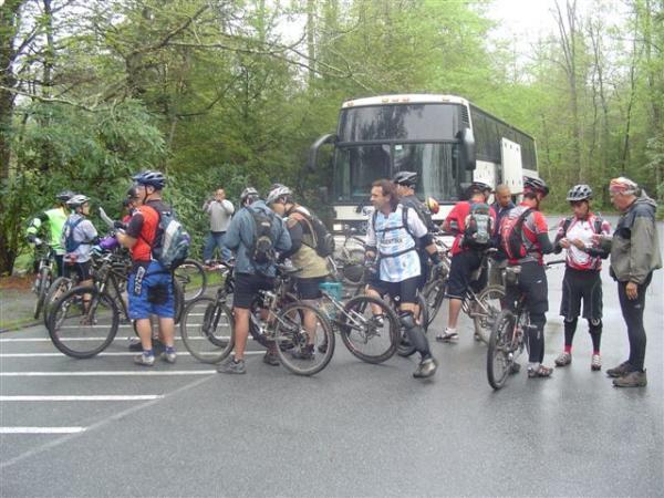 A group of mountain bikers gathered near a bus on a rainy day, surrounded by lush greenery. The riders, wearing helmets and sporting various biking gear, are chatting as they prepare for their biking adventure. Some bikes are parked while others are being held by the riders. The bus is parked in the background, indicating a group outing. Black Mountain mountain bike trail.