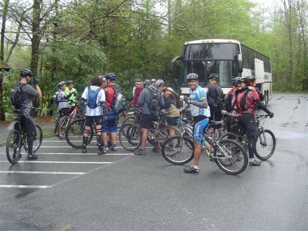 A group of mountain bikers in cycling gear gathered near their bikes in a parking lot, with a large bus parked in the background. The scene is set in a lush, green environment, suggesting an outdoor activity. A few cyclists are holding cameras and water bottles while others appear to be chatting. It is a cloudy day, indicating possible rain. Black Mountain mountain bike trail.