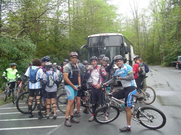 A group of mountain bikers in athletic attire is gathered near a large bus in a forested area. Some riders are around their bikes while others are chatting in the background. The scene depicts a lively atmosphere with greenery around, indicating a recreational biking event. Black Mountain mountain bike trail.