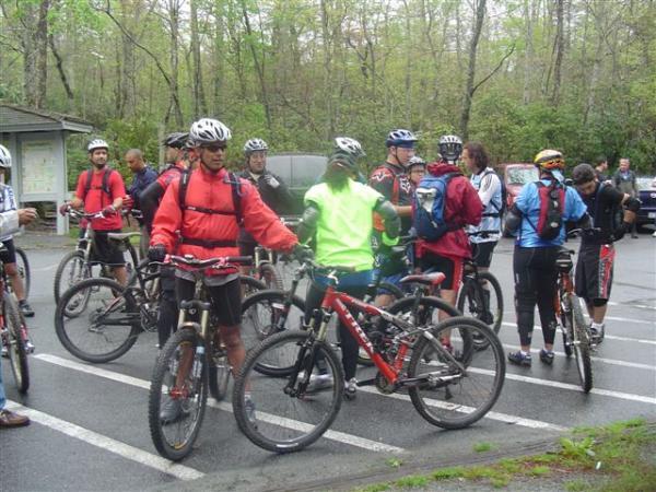 A group of mountain bikers gathered in a parking area, discussing plans and preparing for a ride. They are wearing cycling helmets and various brightly colored jerseys, with bicycles positioned nearby. The background features trees and a building, indicating a natural outdoor setting. Black Mountain mountain bike trail.