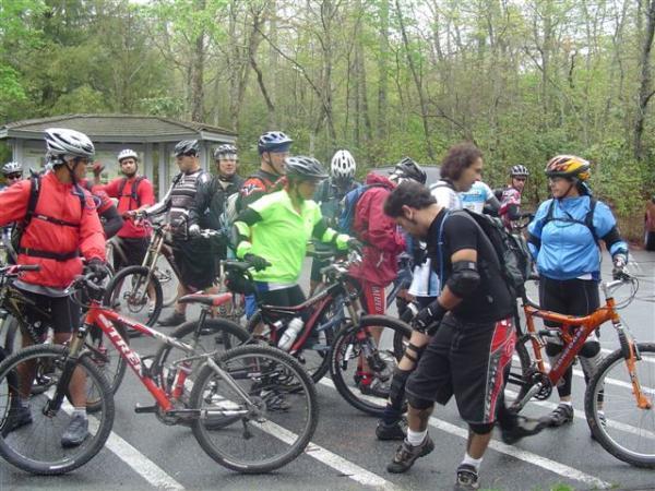 A group of mountain bikers in various colored jerseys and helmets gather in a parking area, surrounded by several mountain bikes. The scene is set in a wooded environment, with trees in the background, indicating an outdoor biking event. Bikers are engaged in conversation and preparing for their ride. Black Mountain mountain bike trail.