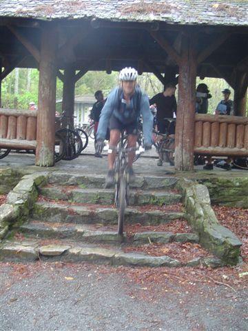 A cyclist in a helmet rides down a set of stone steps, transitioning from a covered pavilion area to a gravel path. In the background, a few other cyclists and individuals can be seen gathered by their bikes. The scene is set in a wooded area with greenery visible. Black Mountain mountain bike trail.